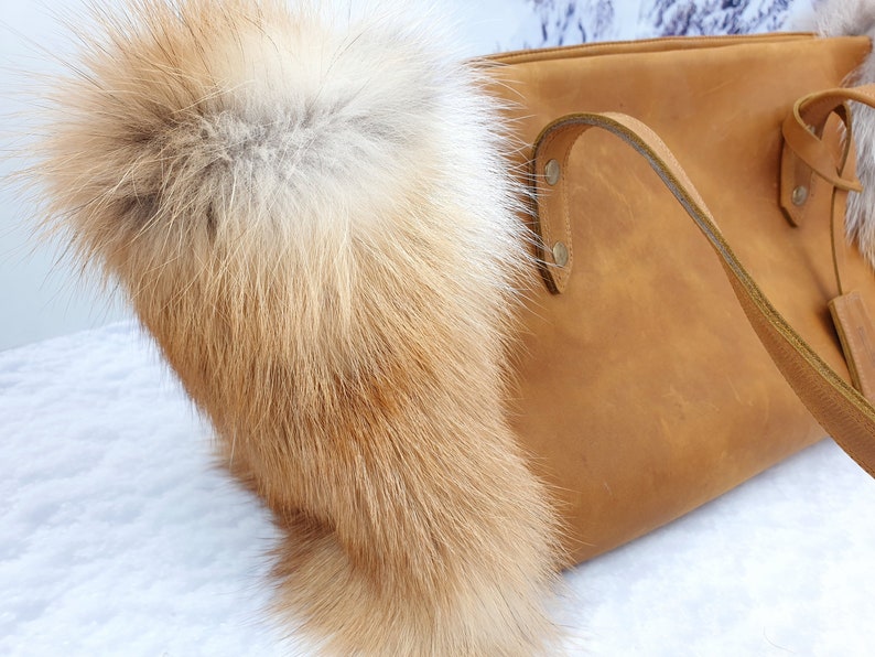 May include: A brown leather tote bag with a fox fur trim. The bag has a long strap and a top zipper closure.