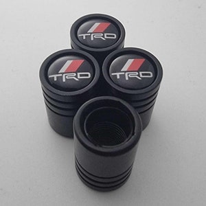 Puede incluir: Conjunto de cuatro tapones de válvula negros con un logotipo TRD rojo y blanco.