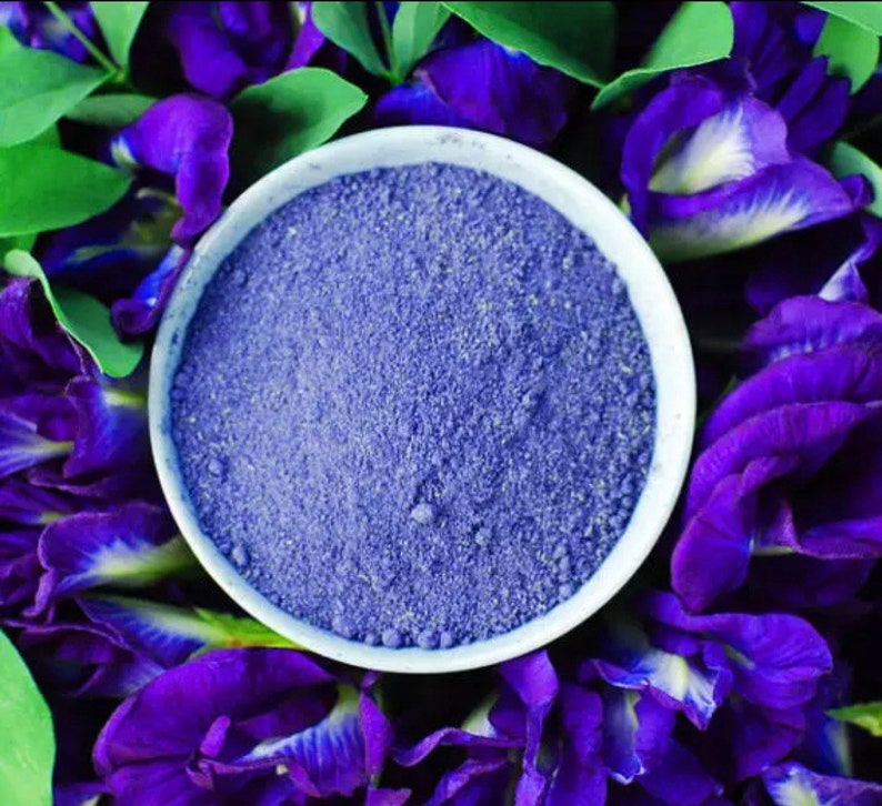 May include: A white bowl filled with finely ground blue powder, surrounded by vibrant purple flowers and green leaves. The powder has a granular texture, contrasting with the smooth petals. The image is a close-up, highlighting the color and texture.