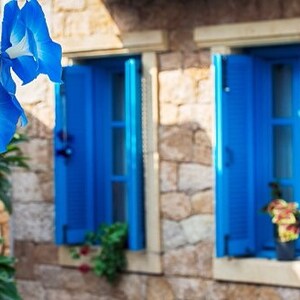May include: A stone building with two blue windows with shutters. The windows are open and there are flowers in front of them.