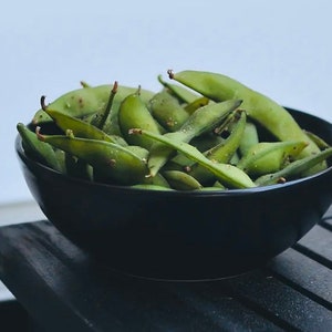 May include: A black bowl filled with green edamame beans. The beans are arranged in a random pattern and some are slightly overlapping. The bowl is sitting on a black wooden surface.