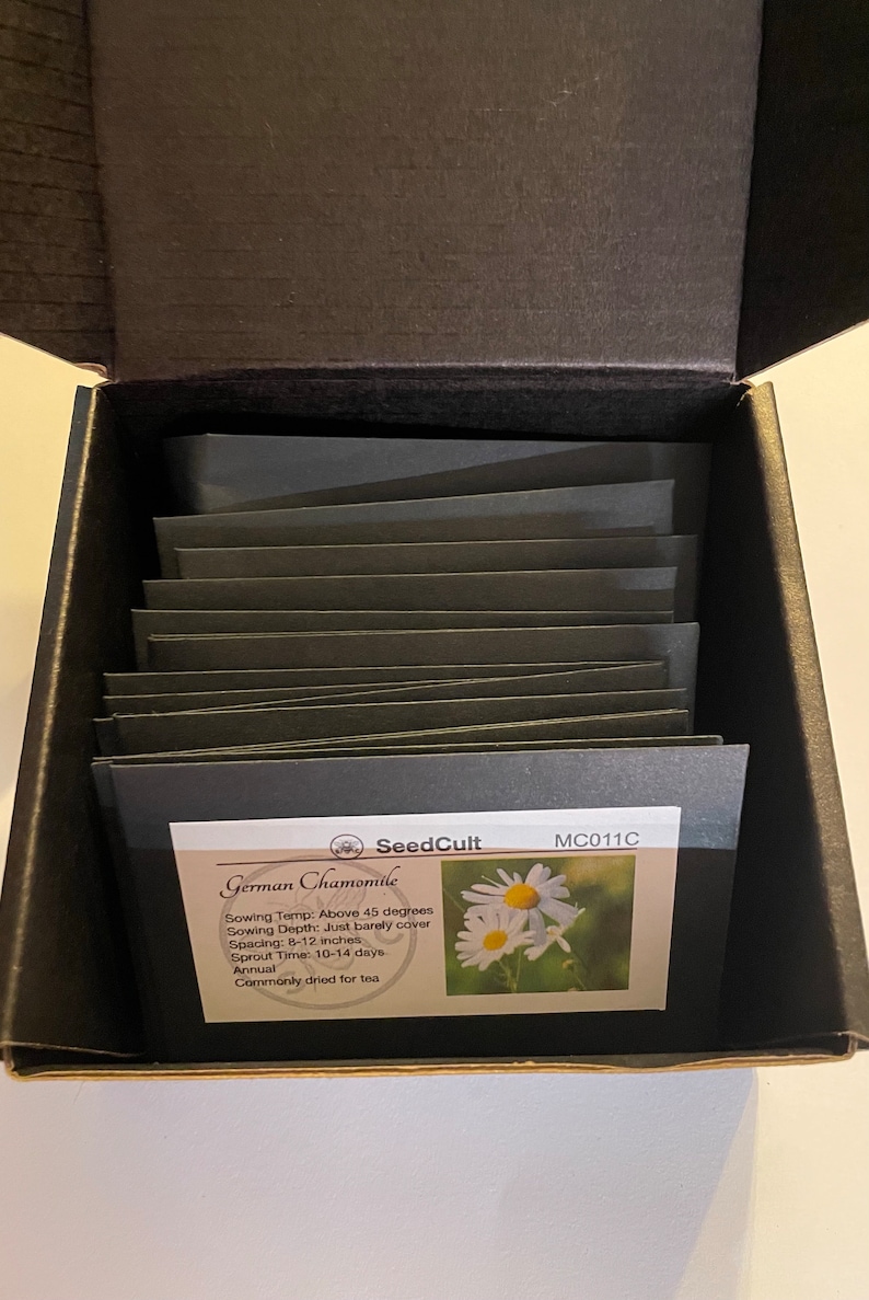 May include: A black box with a white label that says "SeedCult" and "MC011C". The label also says "German Chamomile" and includes information about sowing temperature, depth, spacing, sprout time, and that it is an annual plant commonly dried for tea. The label has a picture of a white daisy.