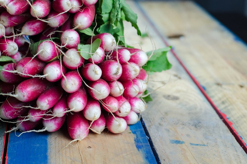 May include: A bunch of fresh radishes with pink and white skin and green leaves, arranged on a wooden surface with blue paint.