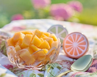 May include: A clear glass bowl filled with diced cantaloupe sits on a floral patterned cloth. Decorative citrus-themed coasters and a gold spoon are also present, suggesting a summer theme.
