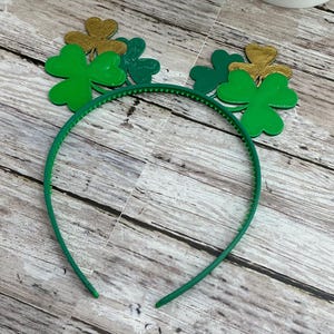 Op de afbeelding: Een groene plastic haarband met klaverdecoraties. De klavers zijn in groen- en goudtinten. De haarband is een accessoire voor St. Patrick's Day.