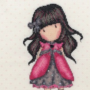 May include: A cross-stitch pattern featuring a girl with long black hair wearing a pink dress with black and white accents. The text "gorjuss" is stitched below the girl.