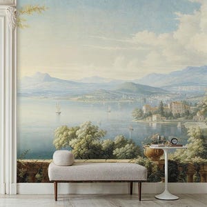 Op de afbeelding: Een muurschildering met een serene meerlandschap met bergen, zeilboten en gebouwen. Een lichtgrijze bank met een rond kussen staat voor de muurschildering. Een kleine witte tafel bevat een decoratieve pot, glazen en een klok.
