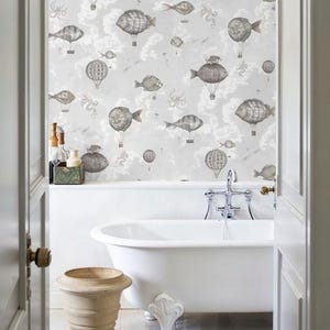 Può includere: Scena di bagno con vasca bianca con piedini a zampa, urna beige e carta da parati con mongolfiere a forma di pesce e nuvole nei toni del grigio e beige. La stanza è incorniciata da una porta.