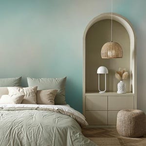 May include: A bedroom interior featuring a bed with sage green bedding and cushions. A woven lampshade hangs above a nightstand with a white lamp and vase. A woven footstool sits on a jute rug.