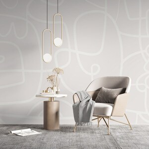 May include: A white armchair with a gold metal frame and a white and black checkered throw blanket sits in a room with a white wall featuring a white abstract design. Two gold pendant lights hang from the ceiling.
