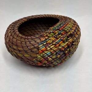 May include: A handcrafted, oval-shaped basket with a woven design. The basket is primarily brown with sections of colorful, textured embellishments in shades of red, yellow, blue, and green. The basket is made of natural materials.