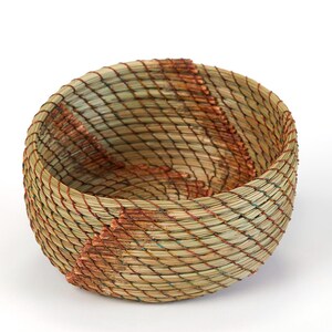 May include: A handwoven basket with a round shape. The basket is made of natural fibers in shades of beige, green, and brown. The weave creates a diamond pattern, with accents of reddish-brown. The basket is empty and sits on a white background.