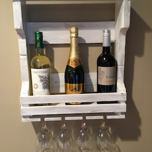 Puede incluir: Un estante de vino de madera blanca con tres botellas de vino y seis copas de vino colgando debajo. Las botellas están etiquetadas con "Caliterra", "St. Martin" y "Champagne".