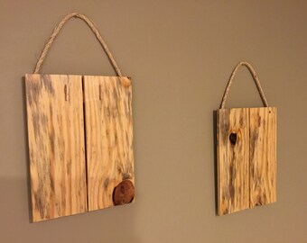 May include: Two rustic wooden signs with rope hangers. The larger sign is made of two pieces of wood and has a knot hole in the bottom right corner. The smaller sign is made of two pieces of wood and has a knot hole in the center.