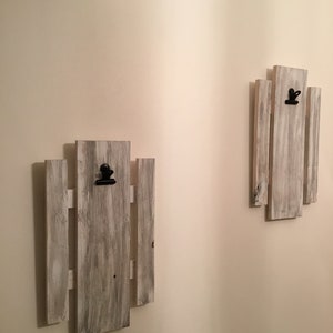 May include: Two whitewashed wooden picture frames with black clips, mounted on a white wall. Each frame is made of vertical slats of wood.