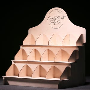 Op de afbeelding: Een houten displaystandaard met meerdere planken om producten te presenteren. De standaard heeft een bord met de tekst "Country Scents Soap Co."