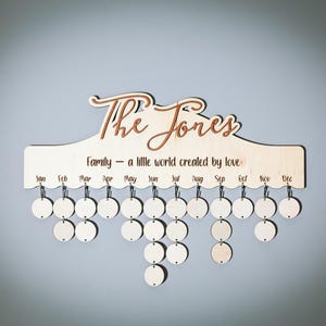 Puede incluir: Un calendario de madera con el texto "The Jones Family - a little world created by love" y los meses del año listados debajo. Hay 12 círculos colgando de cada mes, que representan los días.