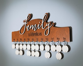 May include: A wooden wall hanging with the word "Family" in cursive script and the word "celebrations" below it. The hanging has 12 hooks with blank circles hanging from each hook, representing the months of the year.