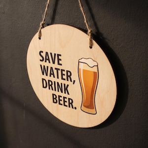 Circular birch plywood sign hanging on a dark wall displaying the quote Save Water Drink Beer alongside a printed beer glass graphic.