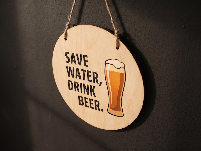 Circular birch plywood sign hanging on a dark wall displaying the quote Save Water Drink Beer alongside a printed beer glass graphic.