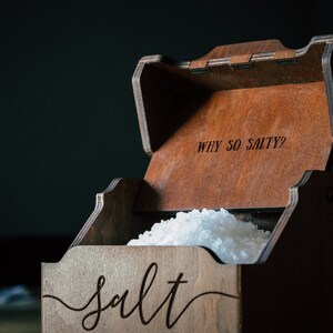 May include: Wooden salt box with a hinged lid, filled with coarse salt. The box is engraved with the word "salt" in a cursive font on the front and "WHY SO SALTY?" on the inside of the lid. The box has a dark brown finish.