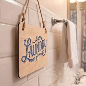 Small indoor door sign reading Laundry in blue script, hanging on white subway tiles in a bright bathroom setting.