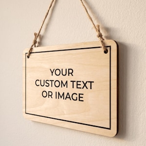 Angled side view of a custom text sign made of thin wood, hanging by natural twine against a plain white wall.