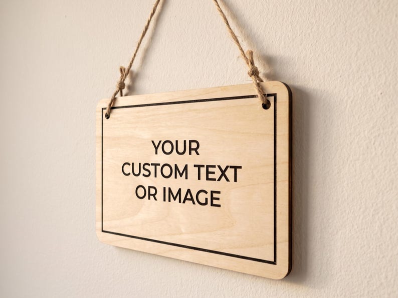 Angled side view of a custom text sign made of thin wood, hanging by natural twine against a plain white wall.