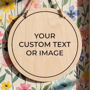 May include: A circular light-colored wooden sign featuring a thin black circular border and the black text "YOUR CUSTOM TEXT OR IMAGE" printed in the center. The sign is suspended by a thin brown twine string attached to the top of the wood.