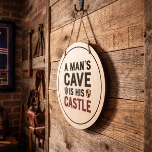Personalized round wood sign hanging on a rustic wooden wall with printed text reading A Man's Cave Is His Castle.