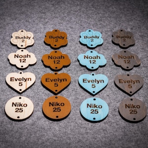 May include: Assortment of wooden pet tags in various shapes and colors, including paw print, cloud, heart, and circle. The tags are engraved with names and numbers, such as "Buddy 2", "Noah 12", "Evelyn 5", and "Niko 25".