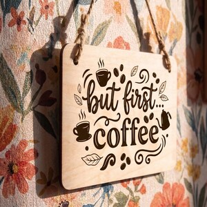 Personalized wood kitchen sign featuring the phrase but first coffee and bean illustrations, hanging on a floral wall.