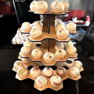 May include: A wooden display stand filled with numerous round bath bombs. The bath bombs are various colors and have labels. The stand has multiple hexagonal shelves, creating a tiered presentation. The background is blurred.