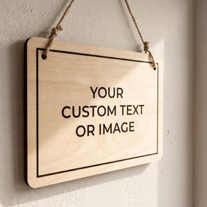 Custom wooden hanging sign with crisp black text on natural birch, illuminated by bright sunlight next to a window.
