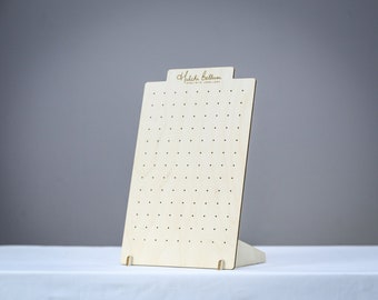 Puede incluir: Un expositor de pendientes de madera con un acabado en madera clara y un fondo blanco. El expositor tiene una base rectangular y un panel vertical con agujeros espaciados uniformemente para sujetar los pendientes. El expositor está etiquetado con "Hulachi Bellum Handmade Jewellery".