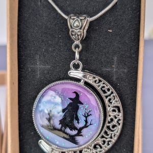May include: Silver-toned pendant necklace featuring a witch silhouette on a branch against a purple and blue background. The pendant is round, framed by a crescent moon design. The necklace is presented in a black box.