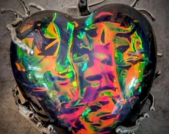 May include: A heart-shaped pendant with a vibrant, iridescent surface displaying a spectrum of colors including pink, green, and orange. The pendant is framed by a silver-colored metal design and has a matching bail for hanging.