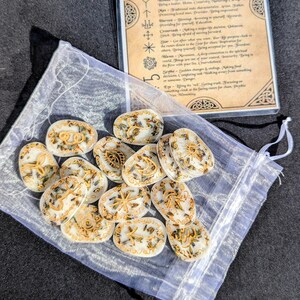 May include: A collection of oval-shaped, white rune stones with gold-colored symbols and botanical accents, presented in a sheer drawstring bag. A card with text and symbols is visible in the background. The runes are for divination.