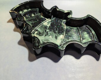 May include: A bat-shaped tray featuring a glossy black outer rim and a dark interior filled with swirling white and green marbled patterns. The surface is embedded with fine glitter particles that catch the light, creating a shimmering effect across the wings and body of the bat-shaped container.