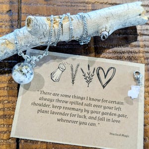 May include: A silver necklace with a clear orb pendant filled with white and green elements, displayed on a wooden branch. A card with text and illustrations is visible. The text reads "Practical Magic".