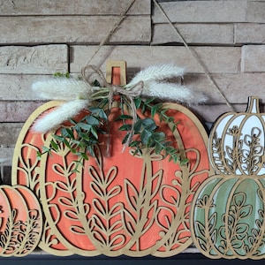 May include: A wooden pumpkin door hanger featuring three pumpkins. The largest pumpkin is orange with a white background and floral designs. The two smaller pumpkins are green and white with floral designs. The pumpkins are hanging from a string.