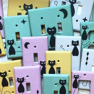 May include: A collection of colorful light switch covers and outlet covers, each hand-painted with a black cat design. The covers are in various pastel colors, including yellow, pink, and teal, with star and moon accents.