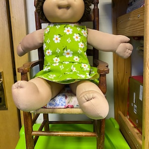 May include: A vintage Cabbage Patch Kid doll with brown yarn hair and a green dress with white daisy print. The doll is seated in a wooden chair with a woven seat, and is resting on a green surface.