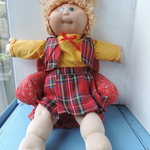 May include: A vintage doll with a soft, round face, blue eyes, and light blonde yarn hair. The doll wears a yellow shirt, a red plaid waistcoat and skirt, and a red bow. The doll is seated in a red floral patterned chair.