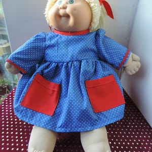 Può includere: Bambola vintage con viso rotondo, occhi blu e capelli biondi. La bambola indossa un vestito blu a pois bianchi e dettagli rossi, tra cui due tasche rosse. Un nastro rosso adorna i capelli della bambola.