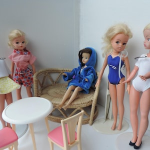 May include: Five dolls, including a doll sitting on a wicker couch, are arranged in a room with a white table and two chairs. The dolls are wearing a variety of outfits, including a white dress, a pink dress, a blue swimsuit, a blue robe, and a white leotard.