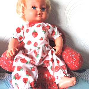 May include: A vintage doll with blonde hair and blue eyes, dressed in a white and red strawberry-patterned outfit. The doll is seated on a red chair with a floral design. The outfit comprises a short-sleeved top and trousers.