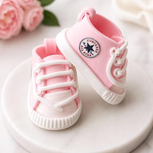 May include: A pair of light pink baby sneakers with white laces, white rubber toe caps, and white soles. The side of the shoe features a circular white patch with the black text Converse All Star and a central black star graphic.