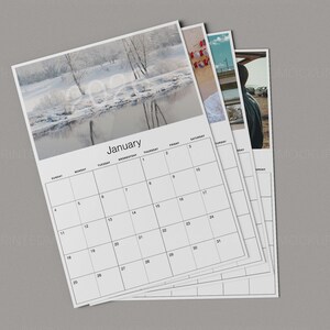 May include: A stack of white paper calendars with a gray background. The top calendar shows the month of January with a winter landscape photo. The calendar grid is black, with the days of the week and dates clearly marked.