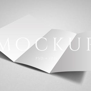 May include: A white, folded brochure mockup with the word "MOCKUP" in large, elegant font. The brochure is open, displaying multiple panels, and is set against a soft gray background. The text "PSD + JPG" is also visible.